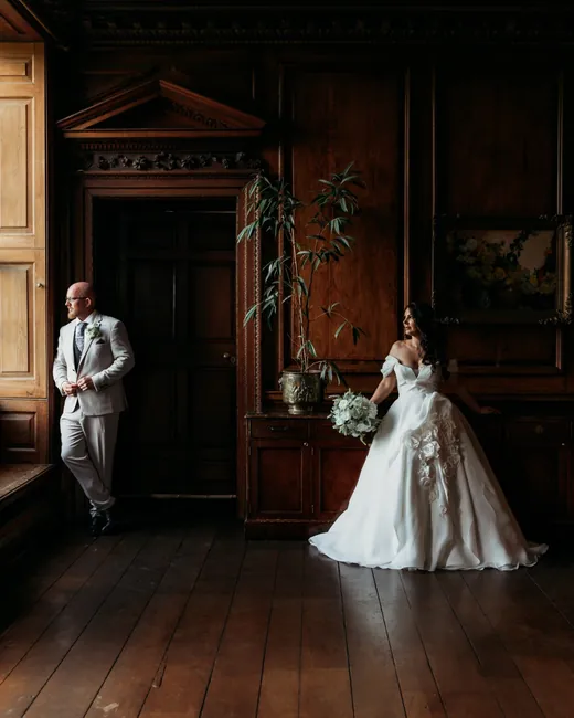 A couple standing apart in a dark wood-panelled room, light falling across the floorboards