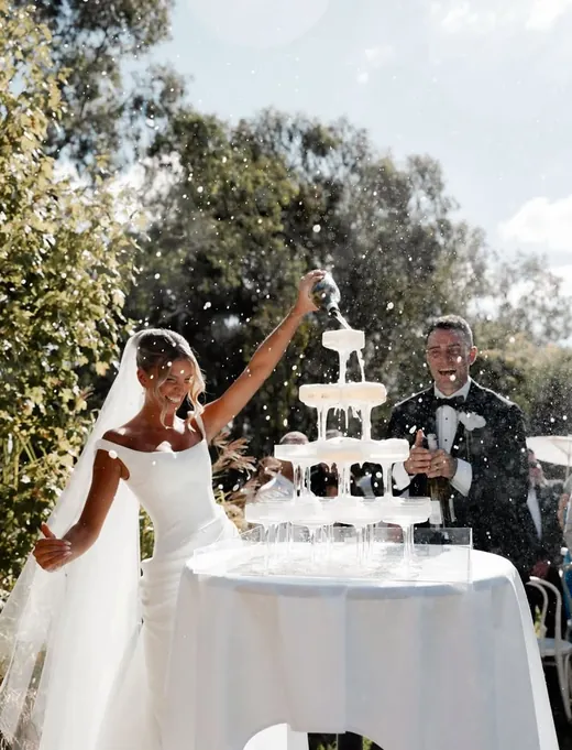 A bride pouring champagne over a coupe tower in a garden while the groom laughs