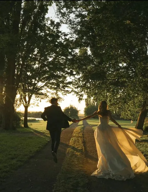 A couple running hand in hand down a tree-lined avenue at sunset, her dress catching the light